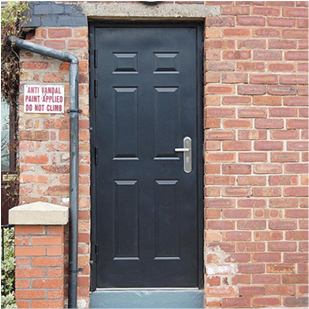 A six panel security door in black on the outside of a brick building with black pipe to the left of it